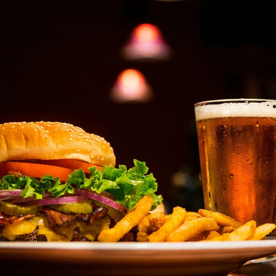 Cheeseburger with lettuce, tomato, pickles, onions, and bacon served with fries beside a pint of beer in a dim restaurant.