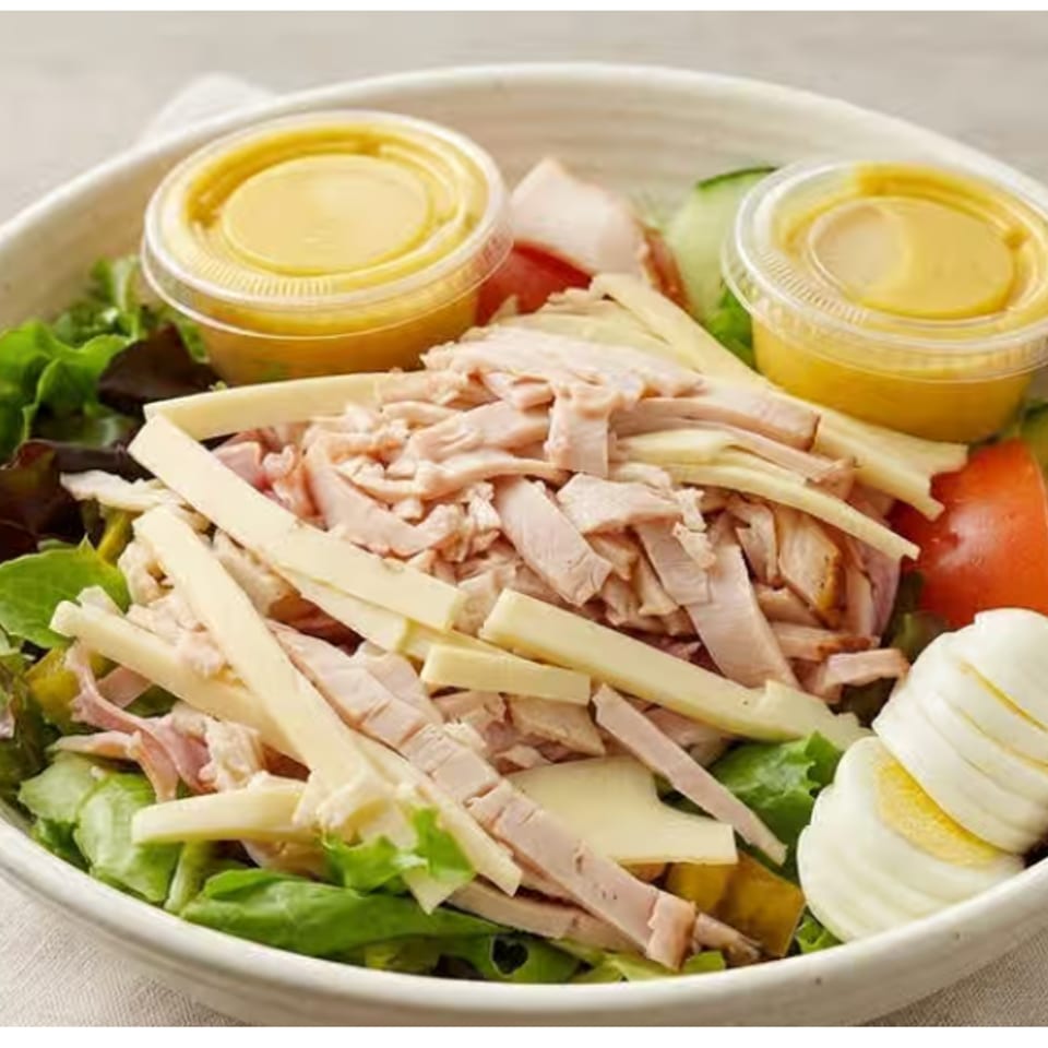 Bowl of mixed greens topped with sliced turkey, bamboo shoots, cucumber, tomato, boiled egg, and two cups of dressing.