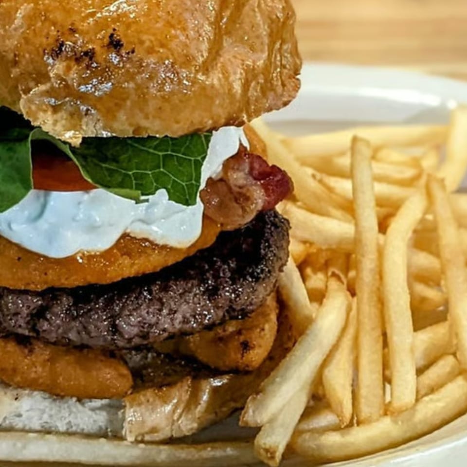 Close-up of a gourmet burger with lettuce, tomato, bacon, sauce, and onion rings beside golden French fries.