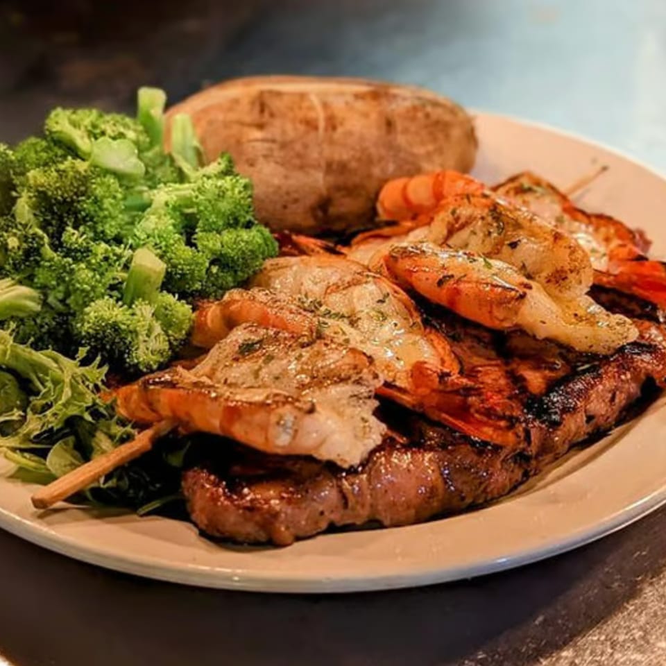Grilled meat and shrimp skewers served with broccoli, leafy greens, carrots, and a baked potato on a plate.