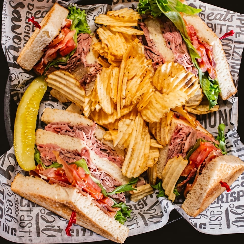 Overhead view of roast beef sandwiches with lettuce and tomatoes, crinkle-cut fries, and a pickle on branded paper.