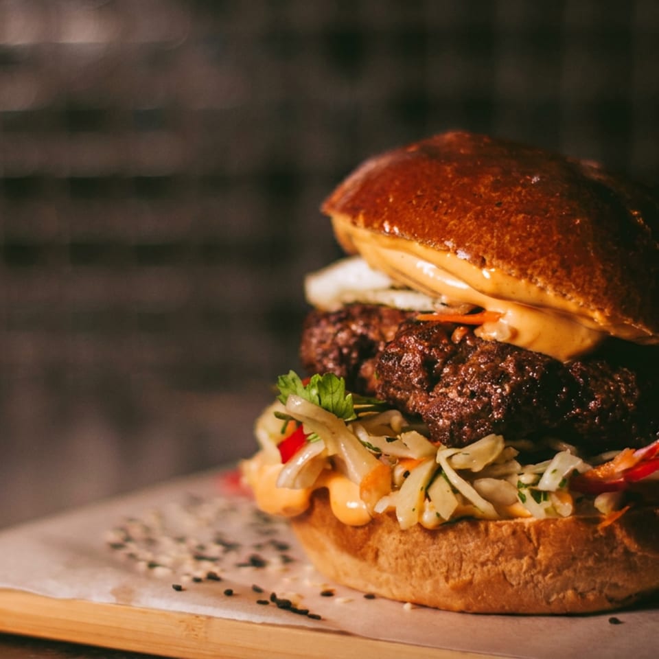 Gourmet burger with two patties, coleslaw, and sauce on a wooden board with sesame seeds.