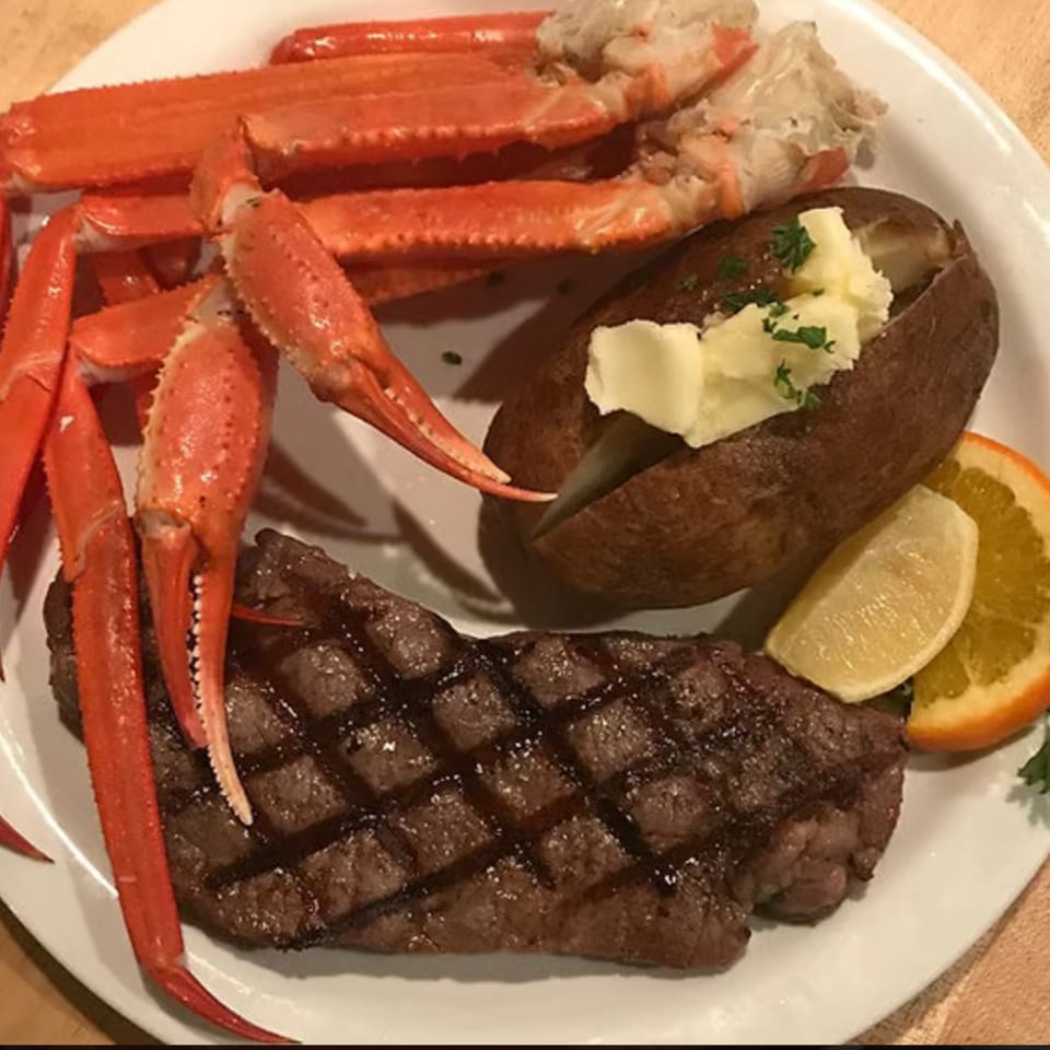 Grilled steak served with red crab legs, a baked potato topped with butter, lemon wedges, and parsley.